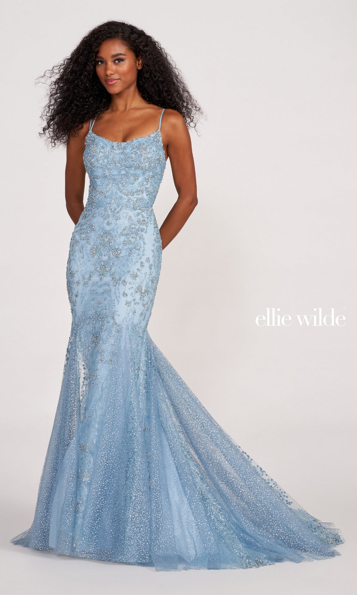 A woman poses against a plain background in the Ellie Wilde Glitter-Jersey Long Mermaid Prom Gown, featuring a light blue fitted silhouette with sequin embellishments and thin straps.